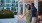 A man and a woman hold hands and walk outside next to chairs and a flower bed